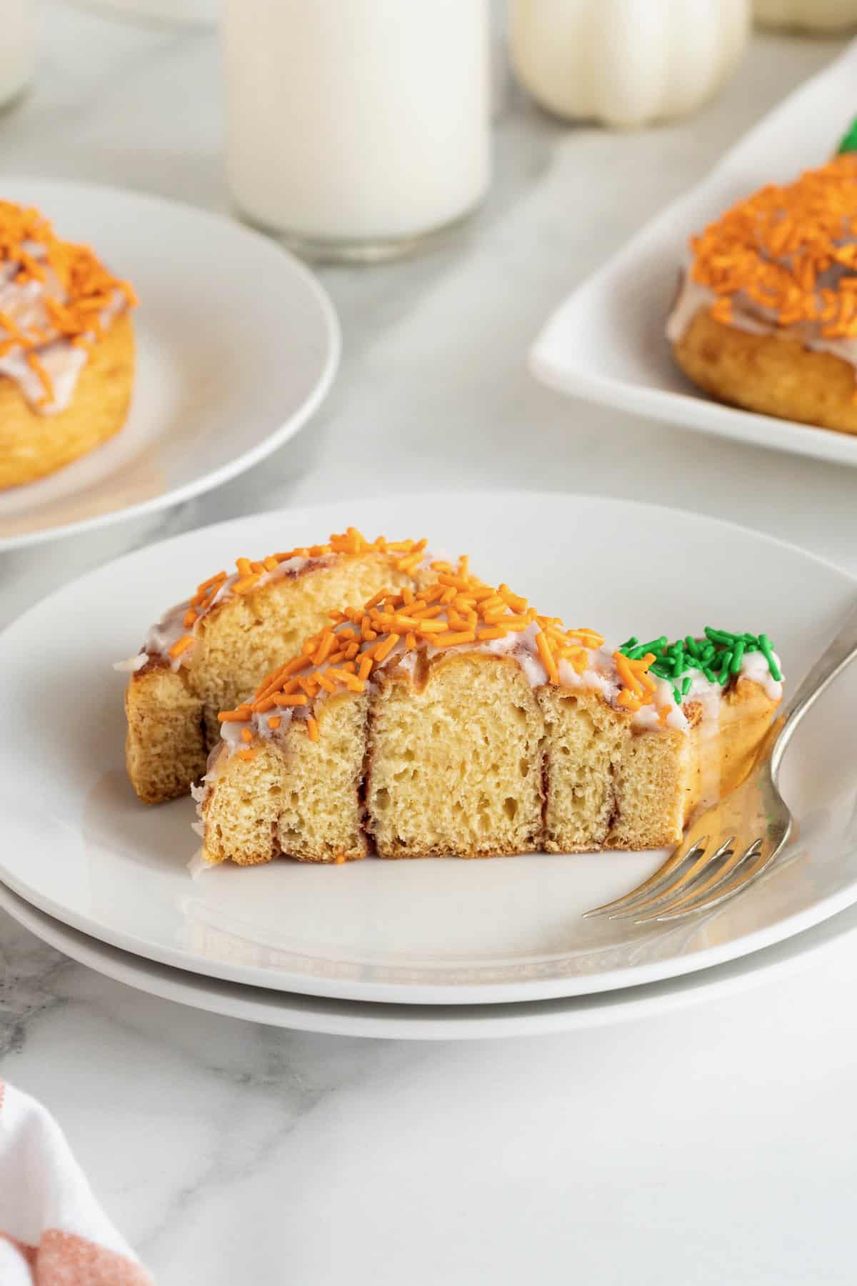 A pumpkin shaped cinnamon roll cut in half with a fork resting beside it on a small white plate.