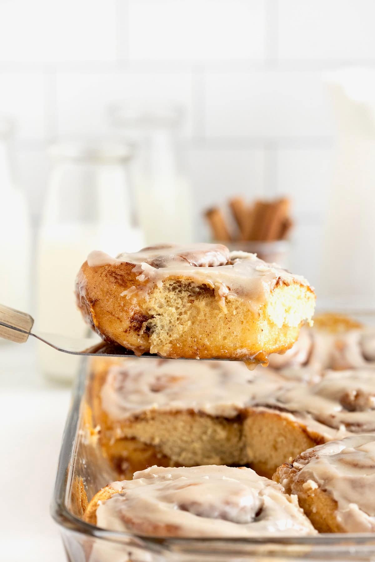 A cinnamon roll on a metal spatula above a glass baking dish of cinnamon rolls.