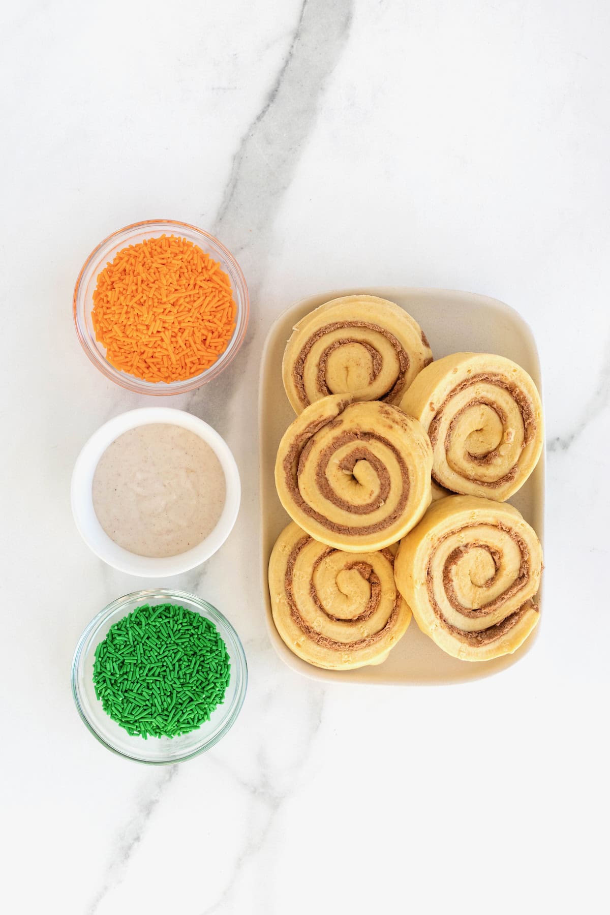 Ingredients to make pumpkin shaped cinnamon rolls in white dishes on a white marble counter.