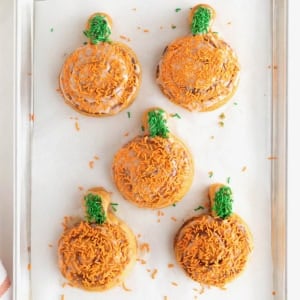 Five frosted pumpkin shaped cinnamon rolls with orange and green sprinkles on a parchment lined aluminum baking sheet.