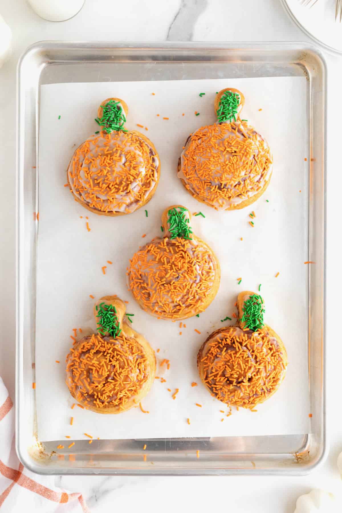 Five frosted pumpkin shaped cinnamon rolls with orange and green sprinkles on a parchment lined aluminum baking sheet.