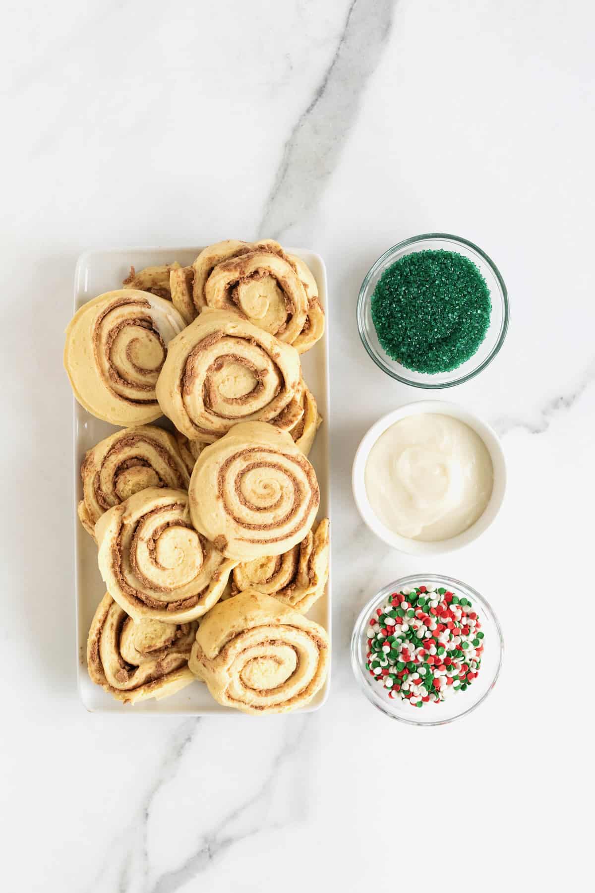 Ingredients to make a Christmas tree cinnamon roll in white dishes on a white marble counter.