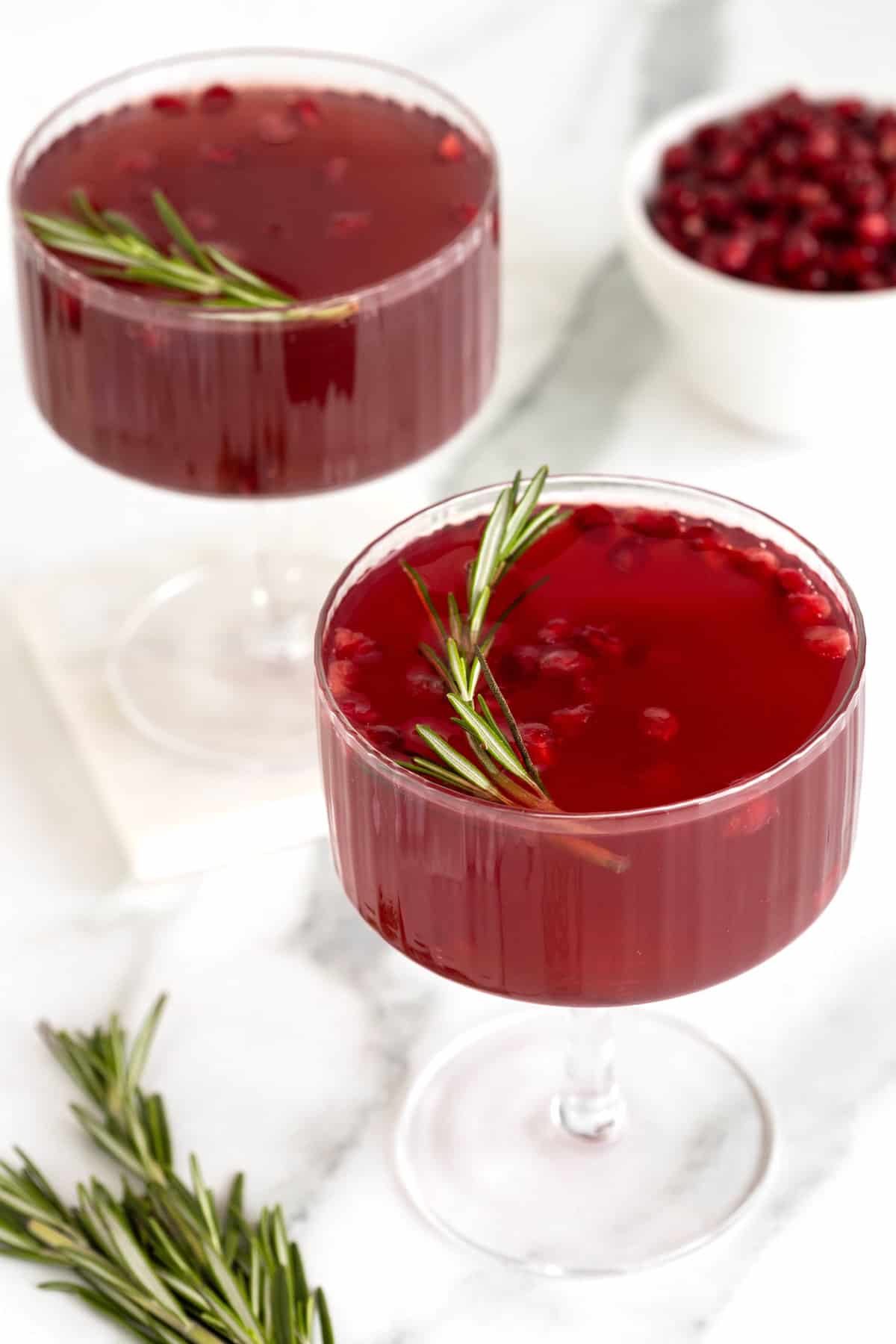 Two dark red cocktails in stemmed coupe glasses with pieces of pomegranate and a sprig of rosemary floating in them.