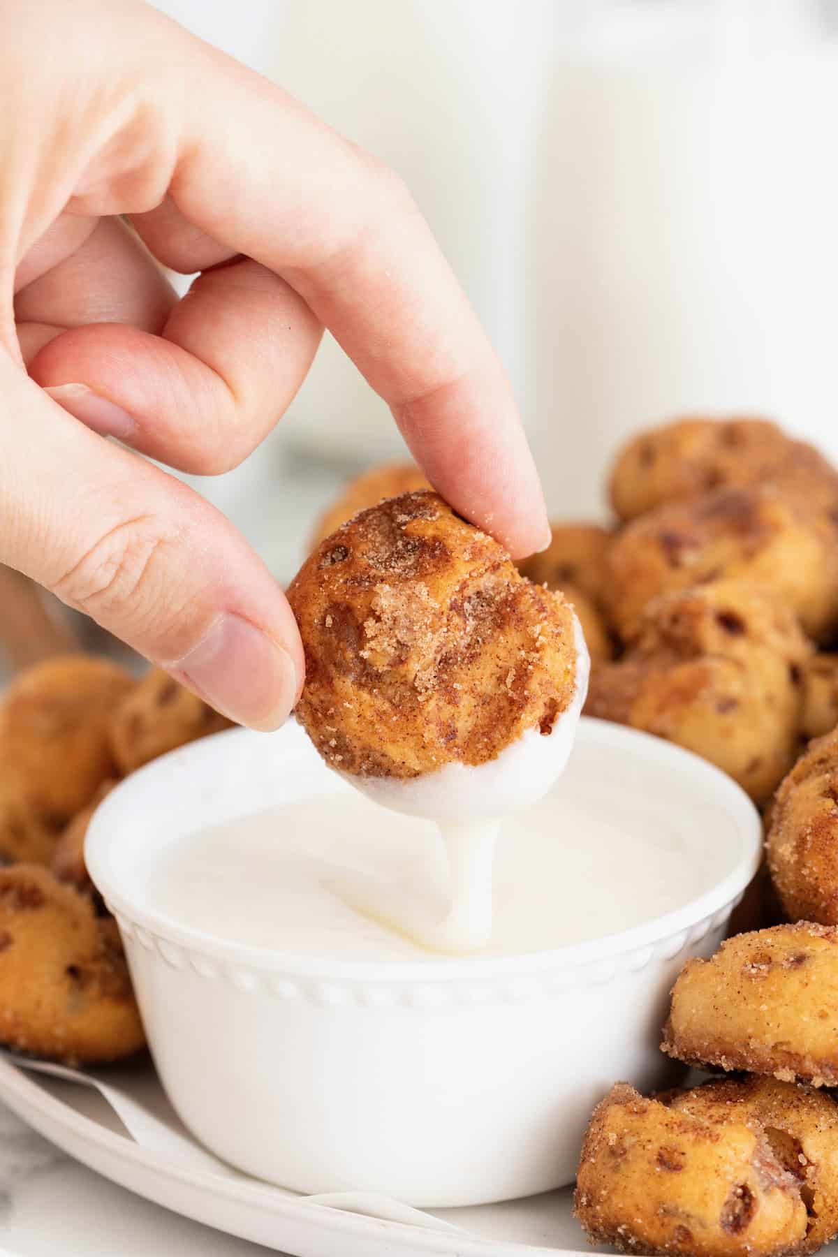 A hand holding a cinnamon roll bite between thumb and forefinger, dipping it in icing. 