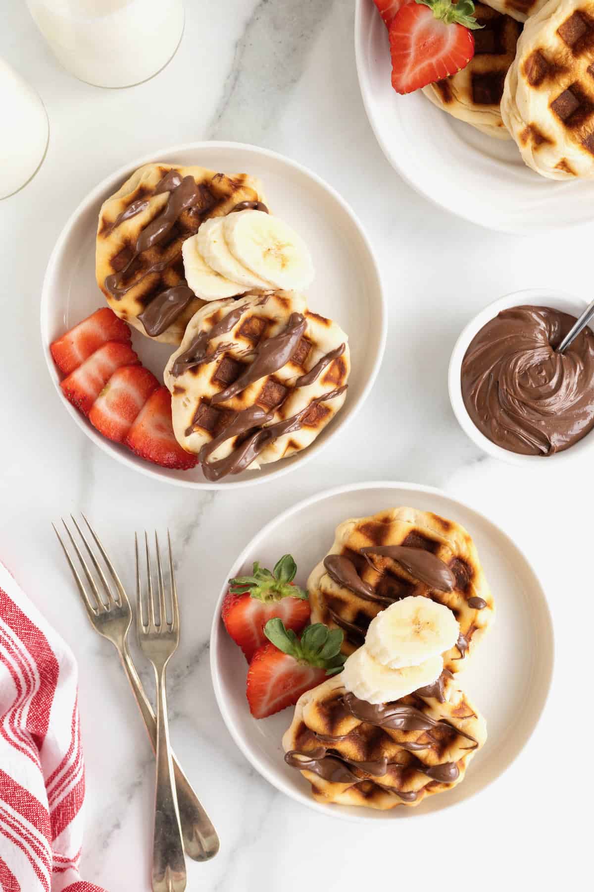 Two white rimmed plates each with two Nutella waffles drizzled with Nutella and topped with banana slices.