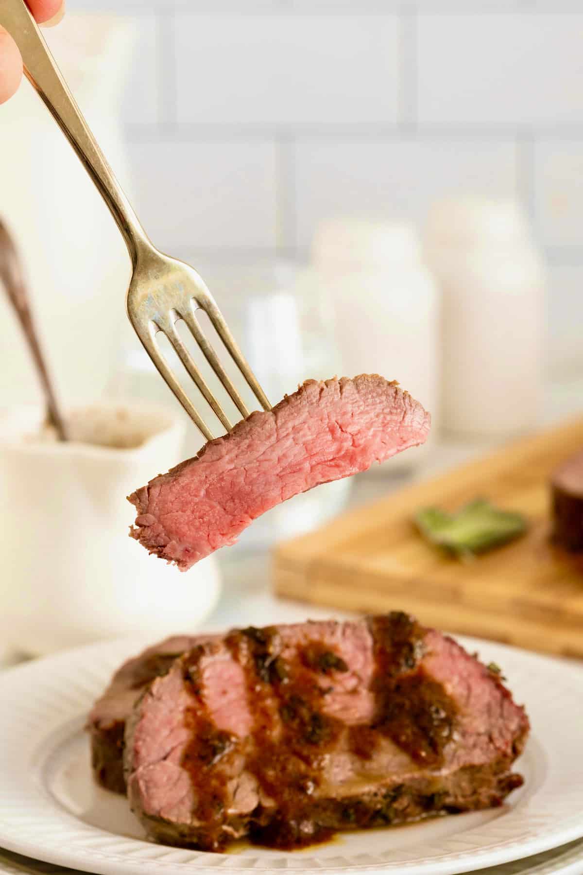 A piece of beef tenderloin on a fork above a plate of sliced beef tenderloin in pan sauce.