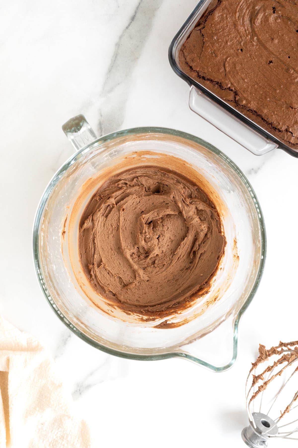 A glass mixing bowl of chocolate frosting on a white marble counter.