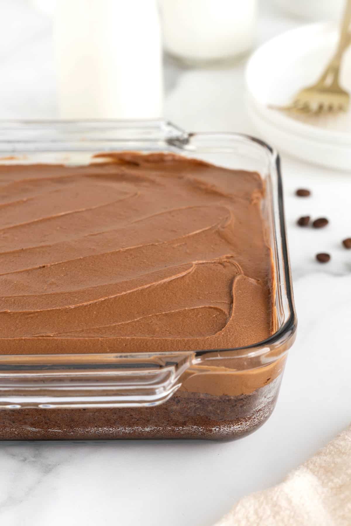 A square glass baking dish of frosted brownies on a white marble counter.