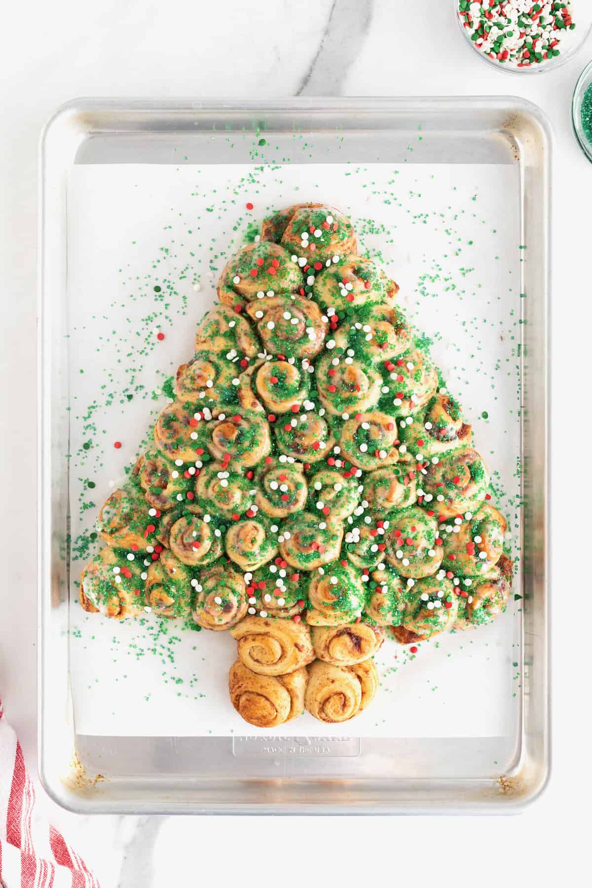 A cinnamon roll Christmas tree sprinkled with green sugar crystals and red and green holiday sprinkles on a parchment lined baking sheet.