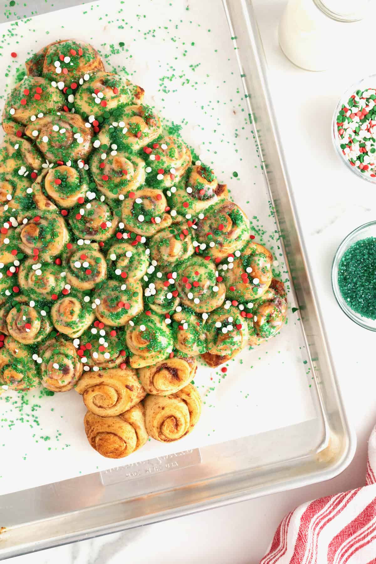 A cinnamon roll Christmas tree sprinkled with green sugar crystals and red and green holiday sprinkles on a parchment lined baking sheet.