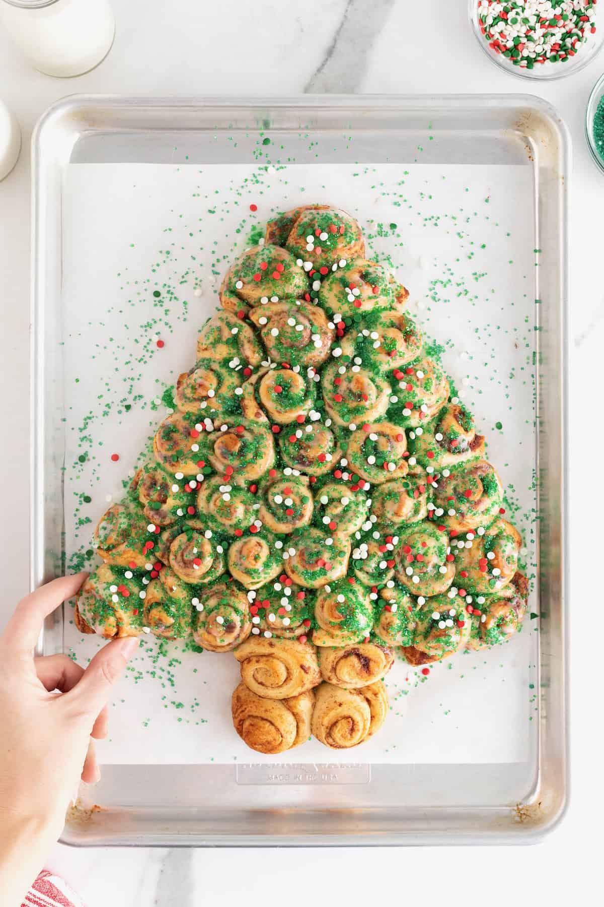 A hand taking a piece of a cinnamon roll pull-apart shaped like a Christmas tree topped with holiday sprinkles.