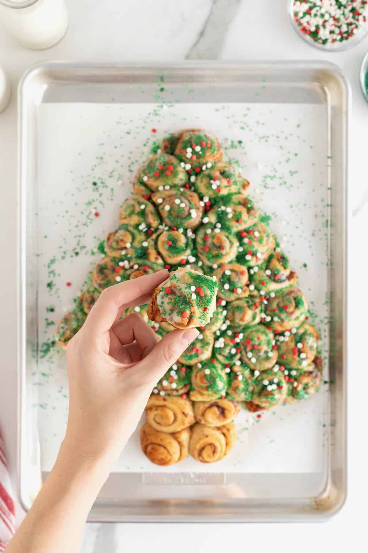 A hand taking a piece of a cinnamon roll pull-apart shaped like a Christmas tree topped with holiday sprinkles.