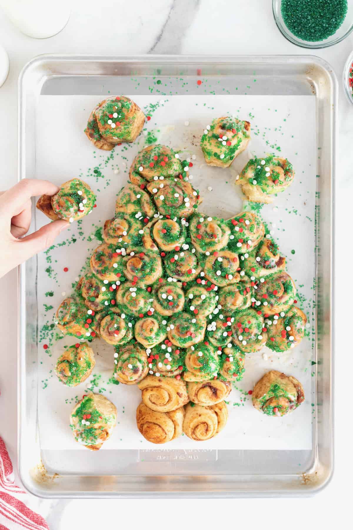 A hand taking a piece of a cinnamon roll pull-apart shaped like a Christmas tree topped with holiday sprinkles.