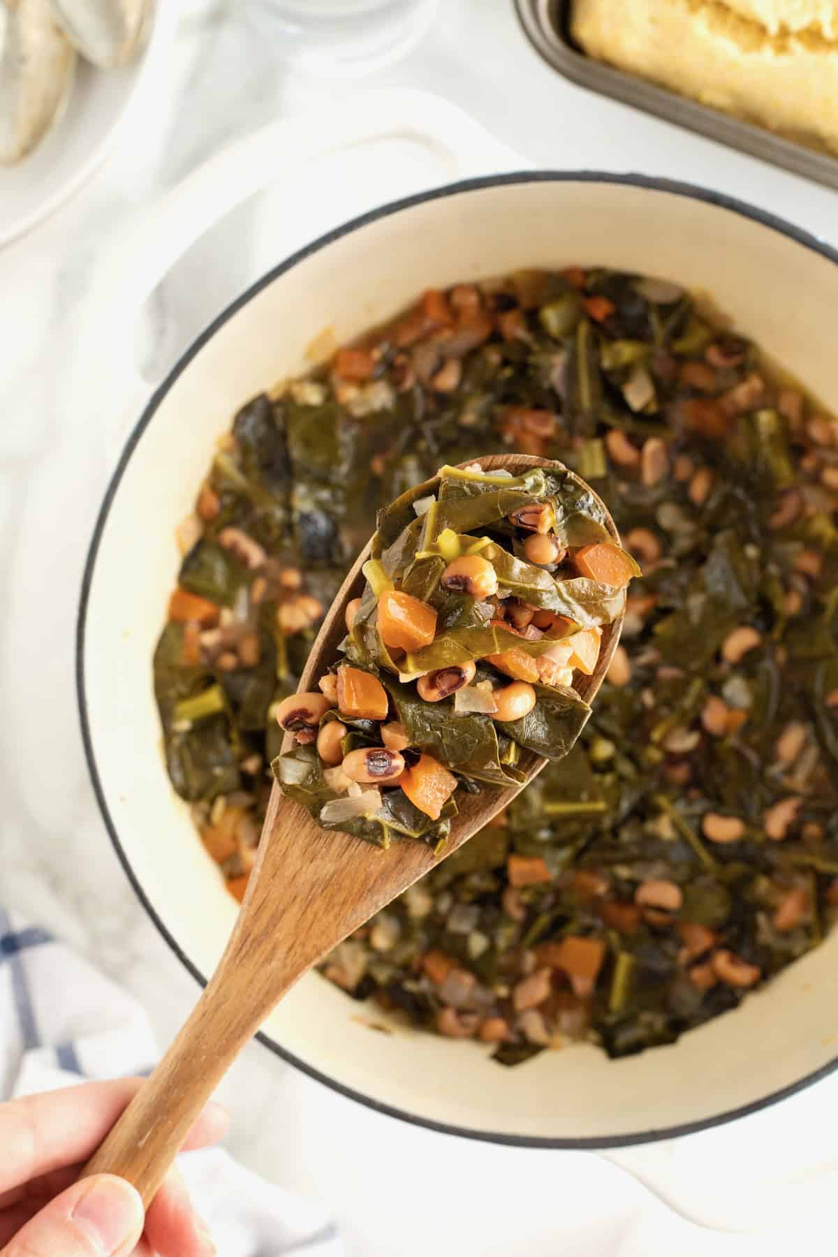 A large white dutch oven filled with black eyed peas and collard greens. A wooden spoon is lifting some of the food from the pot.