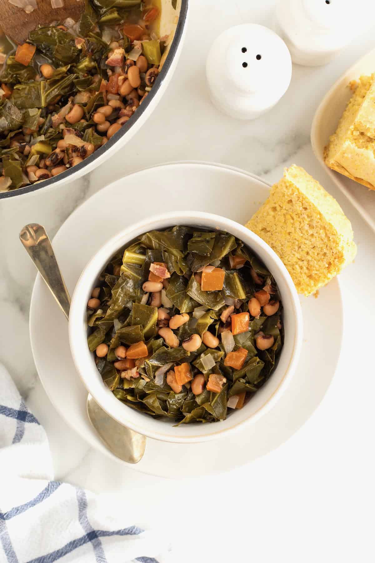 A white soup bowl filled with black eyed peas and collard greens on a white plate with a piece of corn bread and a spoon. 