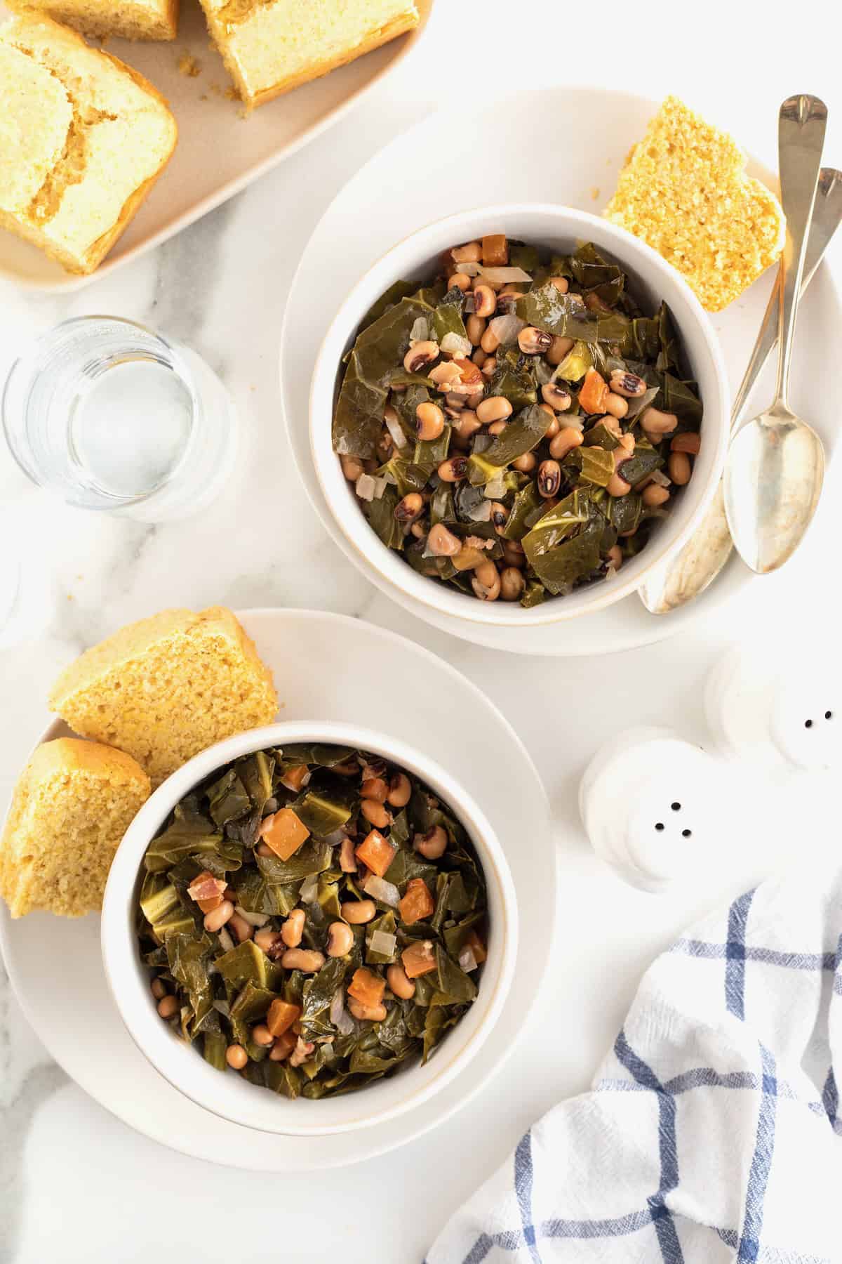 Two bowls of black eye peas on white plates with chunks of cornbread.