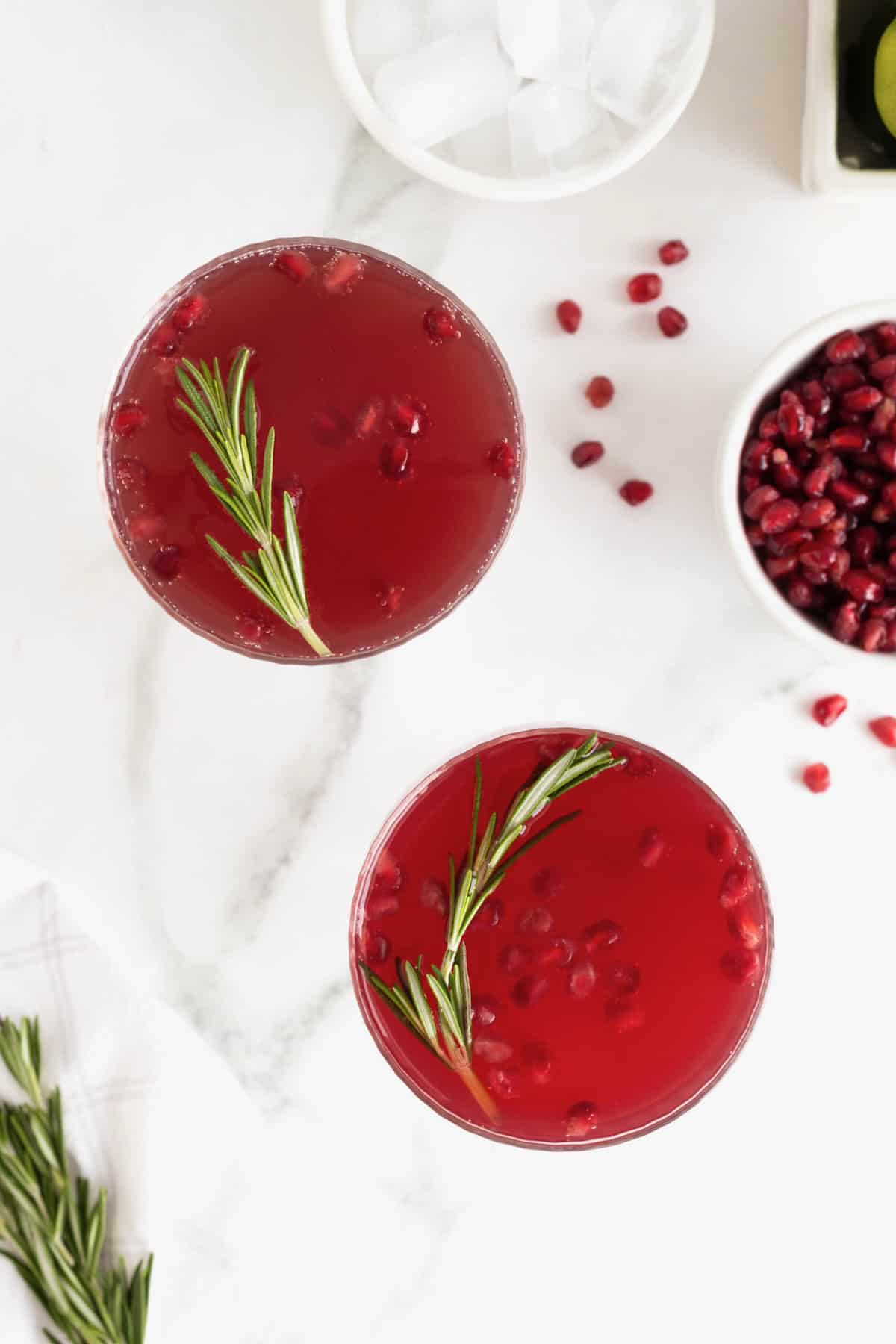 Two dark red cocktails in stemmed coupe glasses with pieces of pomegranate and a sprig of rosemary floating in them.