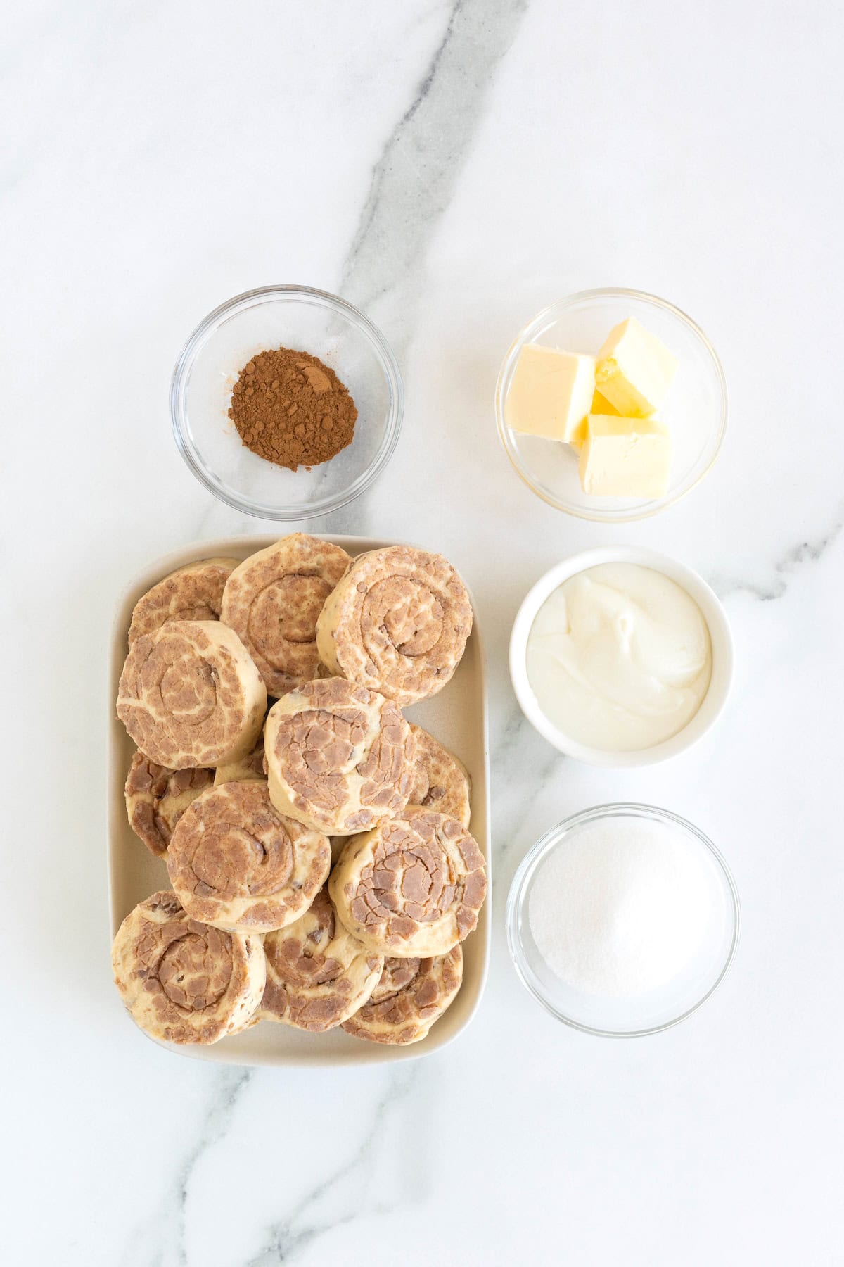 Ingredients to make cinnamon roll bites in glass dishes on a white marble counter.