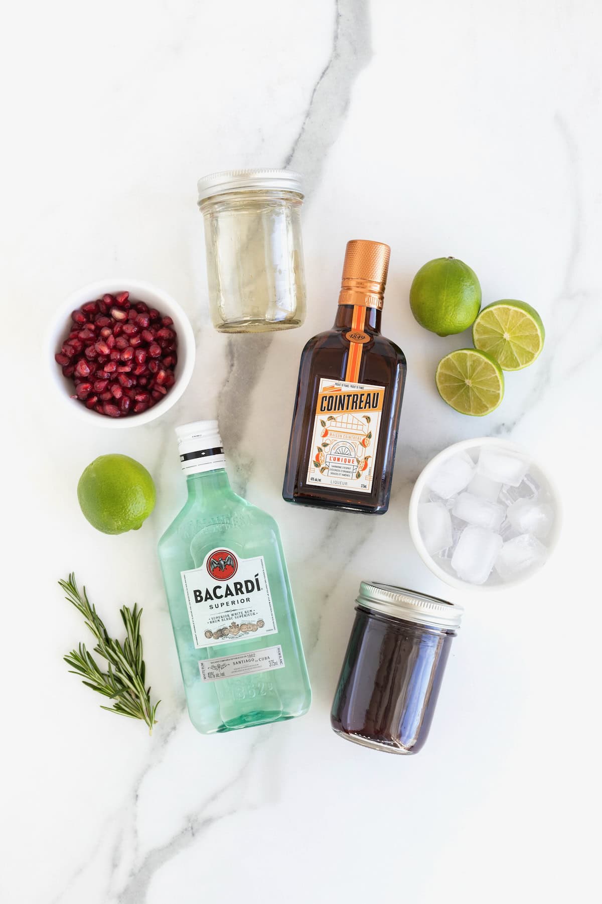 Ingredients to make a pomegranate rosemary holiday cocktail on a white marble counter.