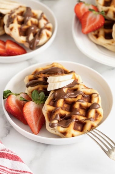 Two round waffles drizzled with Nutella on a small white rimmed plate next to a halved strawberry.