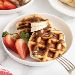 Two round waffles drizzled with Nutella on a small white rimmed plate next to a halved strawberry.