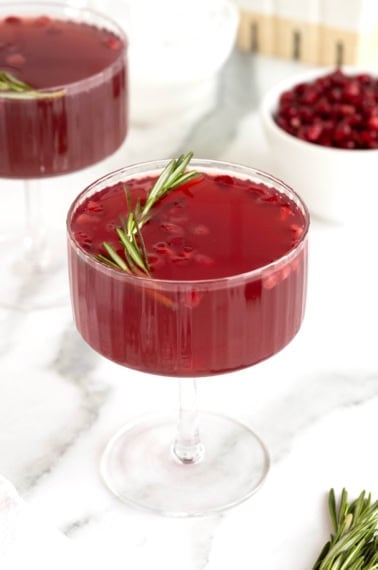 A dark red cocktail in a stemmed glass with pieces of pomegranate and a sprig of rosemary floating in it.