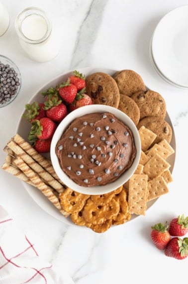 A white bowl of chocolate cheesecake dip topped with chocolate chips and surrounded with various crackers, cookies, pretzels and strawberries.