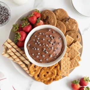 A white bowl of chocolate cheesecake dip topped with chocolate chips and surrounded with various crackers, cookies, pretzels and strawberries.