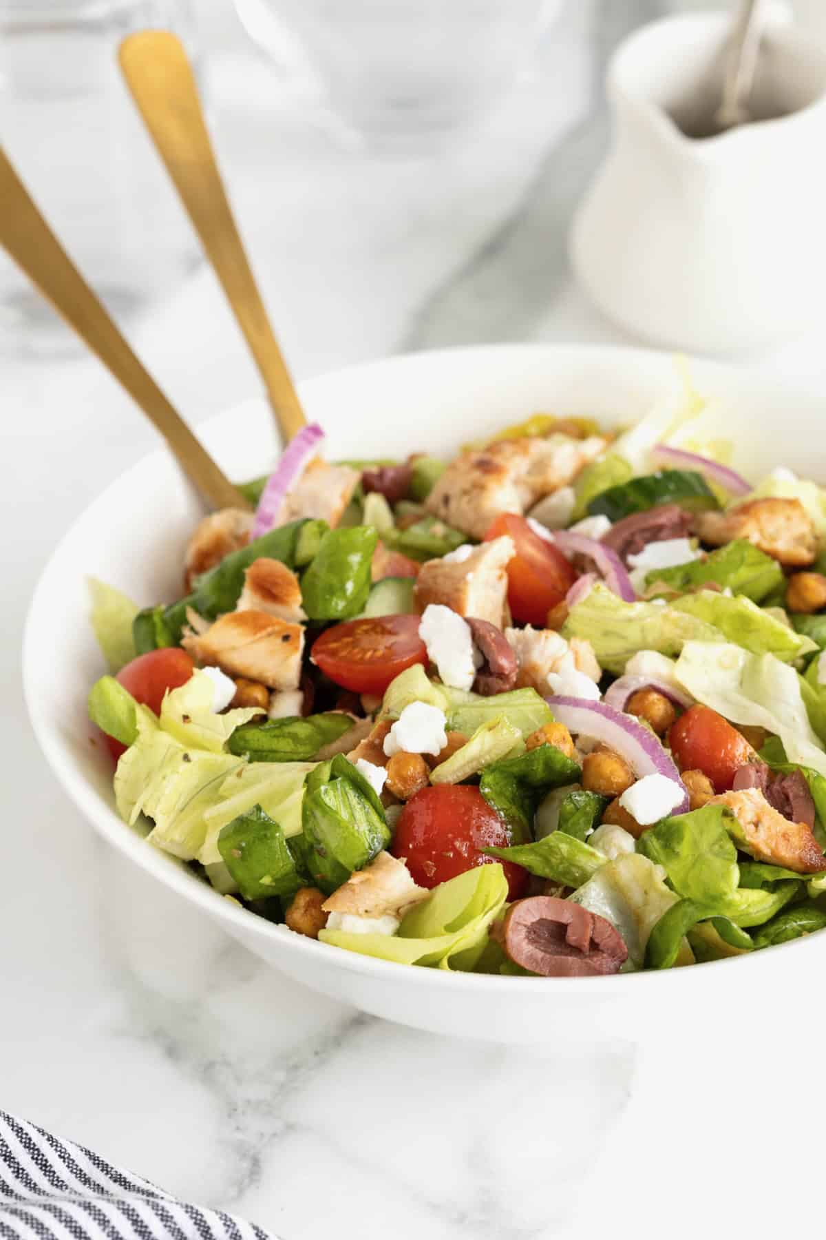 A large white serving bowl with greek salad topped with chicken, tomatoes, sliced greek olives, feta, and roasted chickpeas.