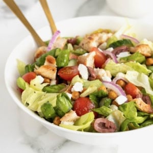 A large white serving bowl with greek salad topped with chicken, tomatoes, sliced greek olives, feta, and roasted chickpeas.