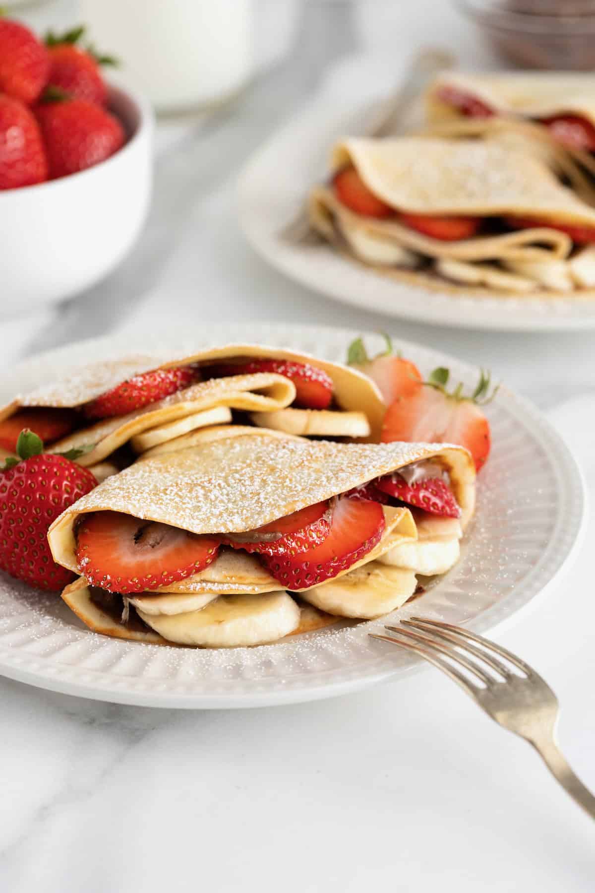 Two crepes on a white fluted plate with a fork resting on the edge of the plate. The crepes are filled with strawberry slices and banana slices and dusted with powdered sugar.