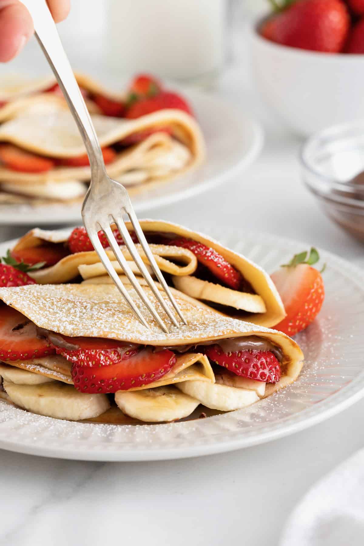 Two crepes on a white fluted plate with a fork. The crepes are filled with strawberry slices and banana slices and dusted with powdered sugar.