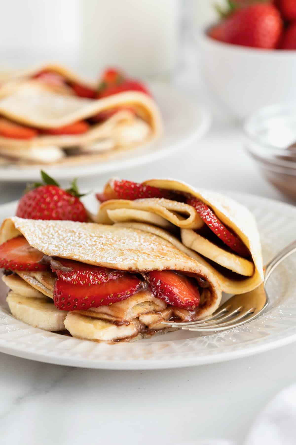 Two crepes on a white fluted plate with a fork. The crepes are filled with strawberry slices and banana slices and dusted with powdered sugar.