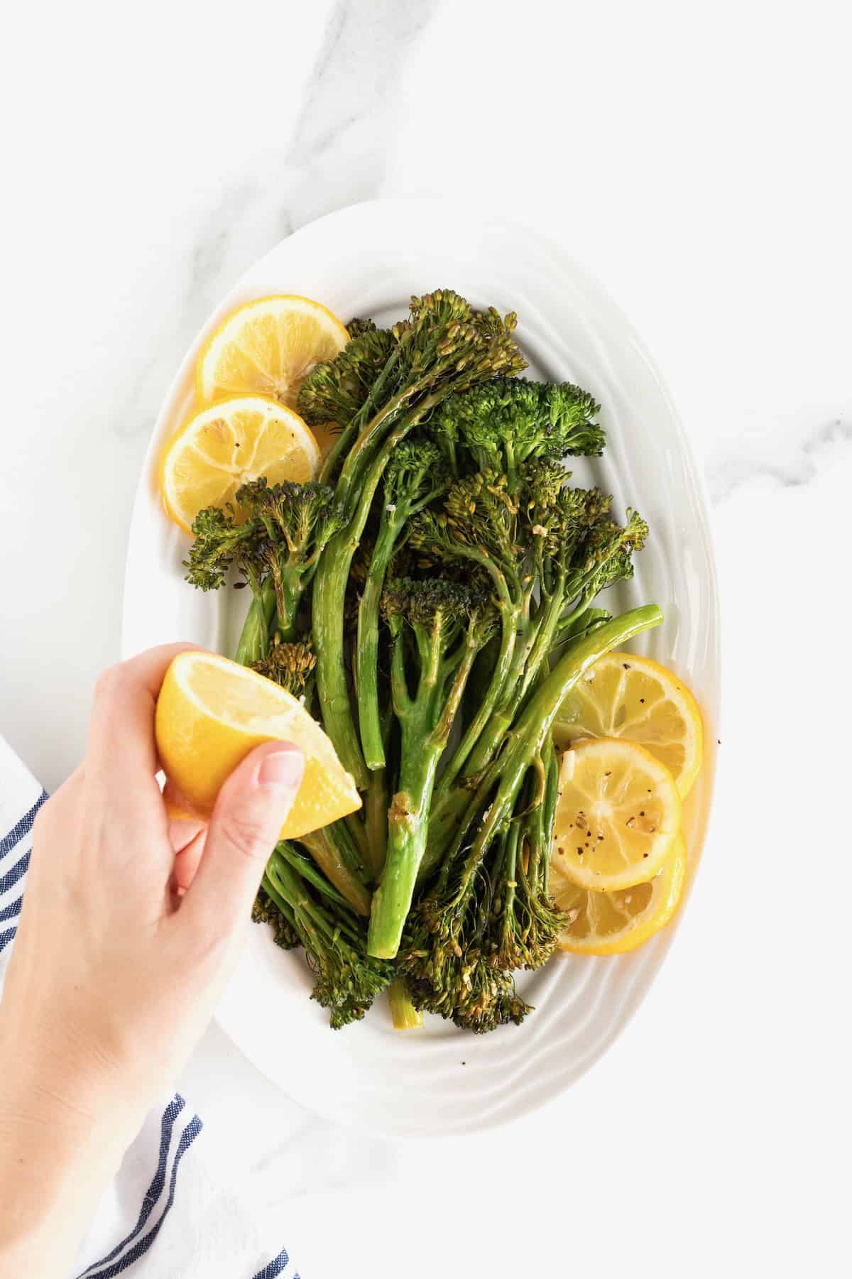 A hand squeezing half of a lemon over a pile of broccolini on a white serving platter.