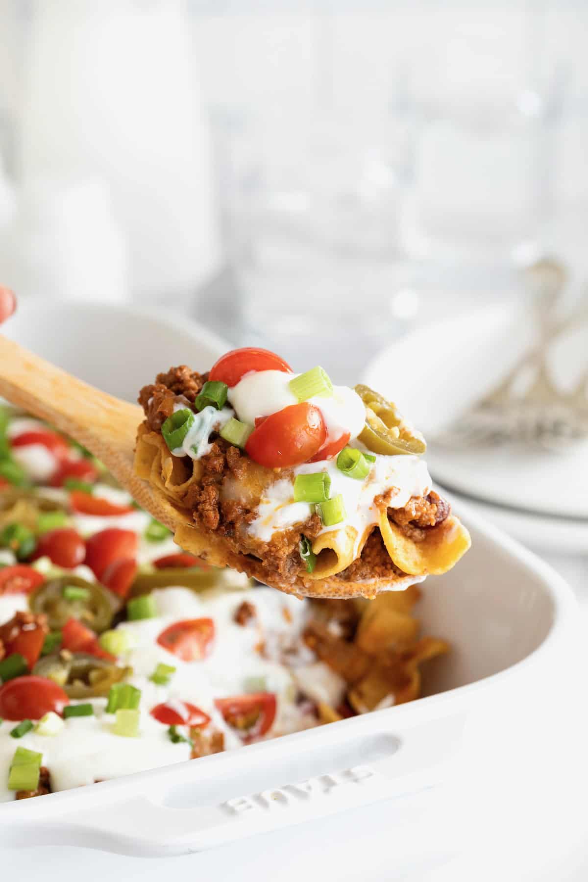 A serving of Frito chili pie casserole on a wooden spoon above a white casserole dish.