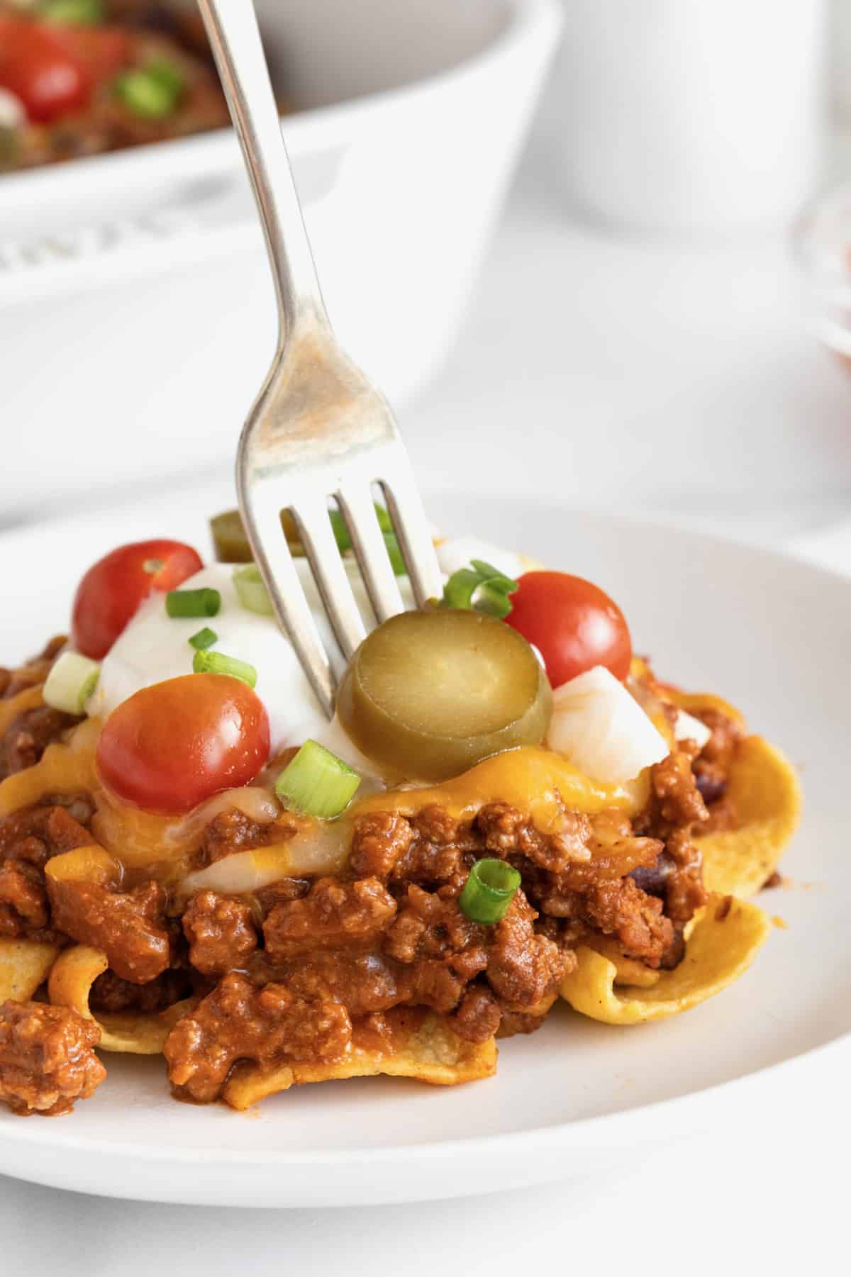 A serving of Frito chili pie casserole on a white plate with a metal fork sticking out.