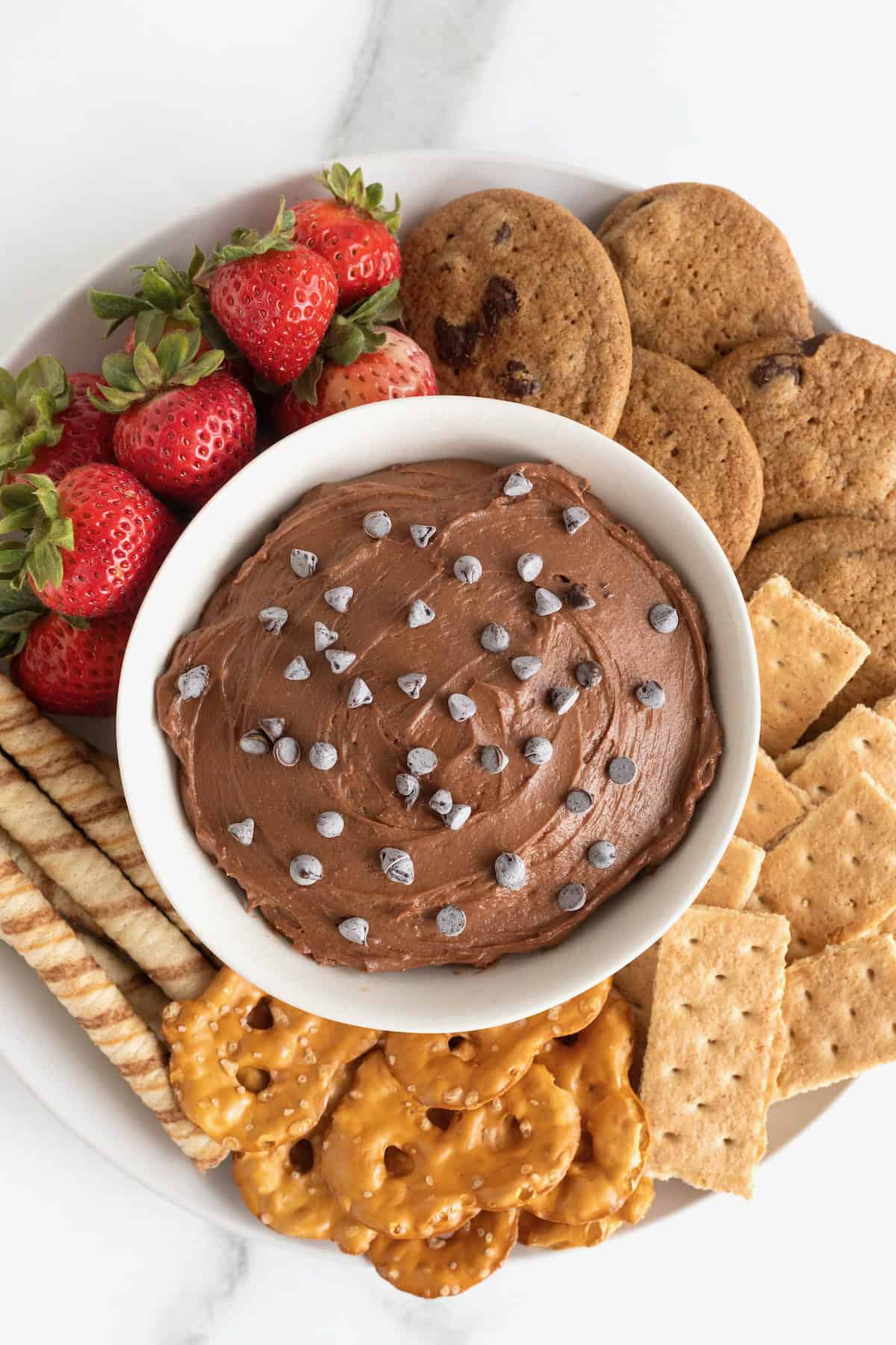 A white bowl of chocolate cheesecake dip topped with chocolate chips and surrounded with various crackers, cookies, pretzels and strawberries.