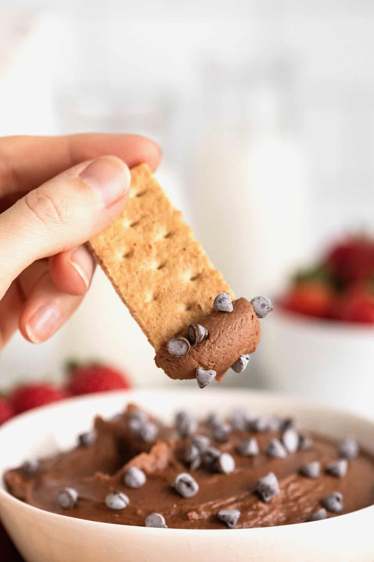 A graham cracker being dipped into chocolate cheesecake dip with mini chocolate chips stuck to it.