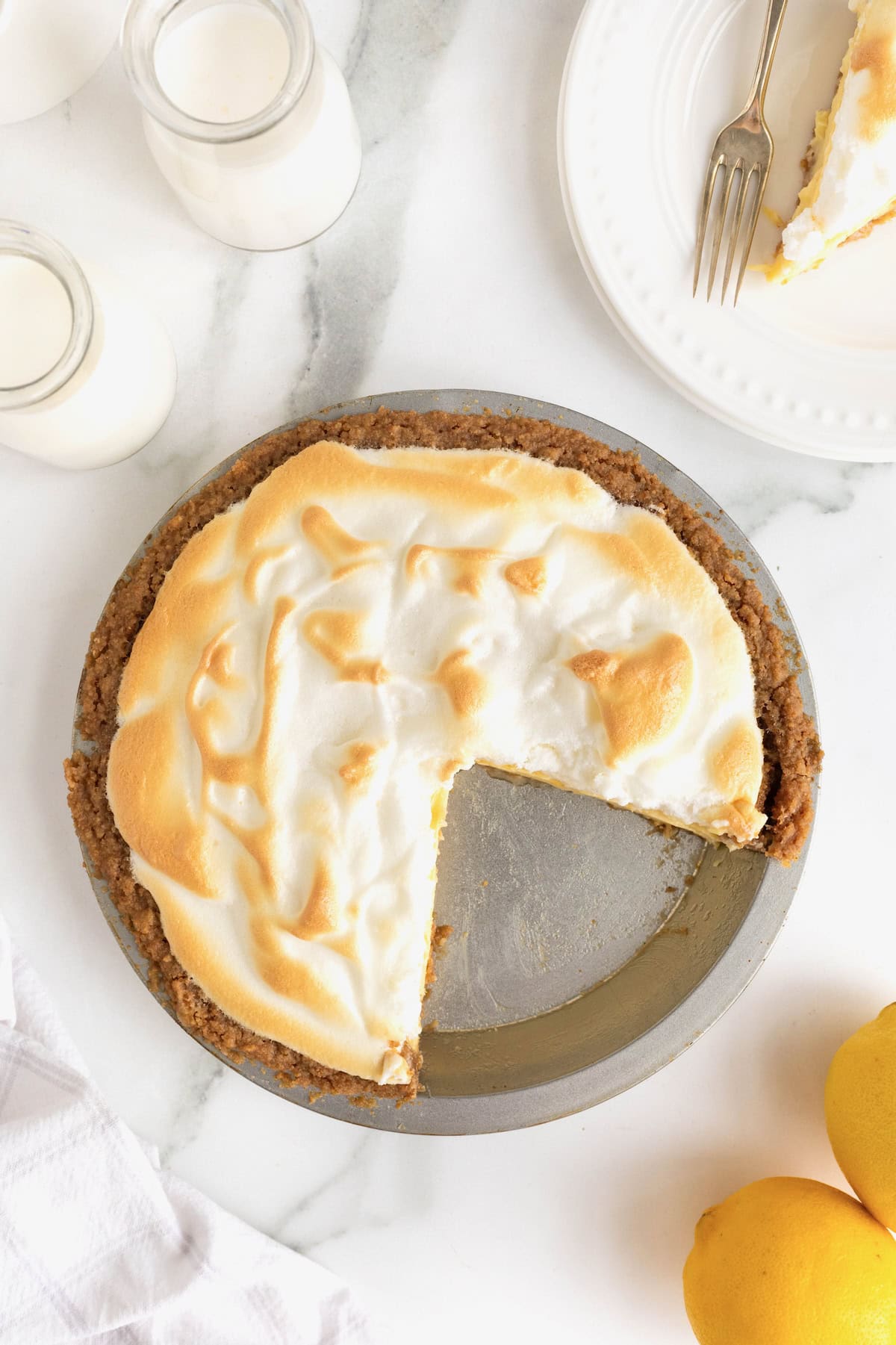 A lemon meringue pie in an aluminum pie plate on a white marble counter. Two slices from the bottom right of the pie have been removed.