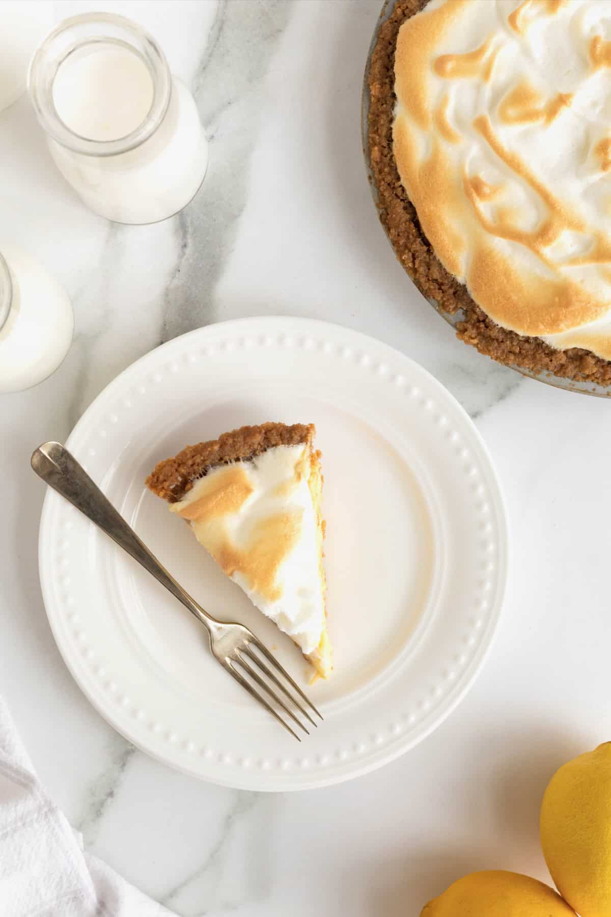 A slice of Lemon Meringue Pie next to a fork on a white dessert plate with beaded details.