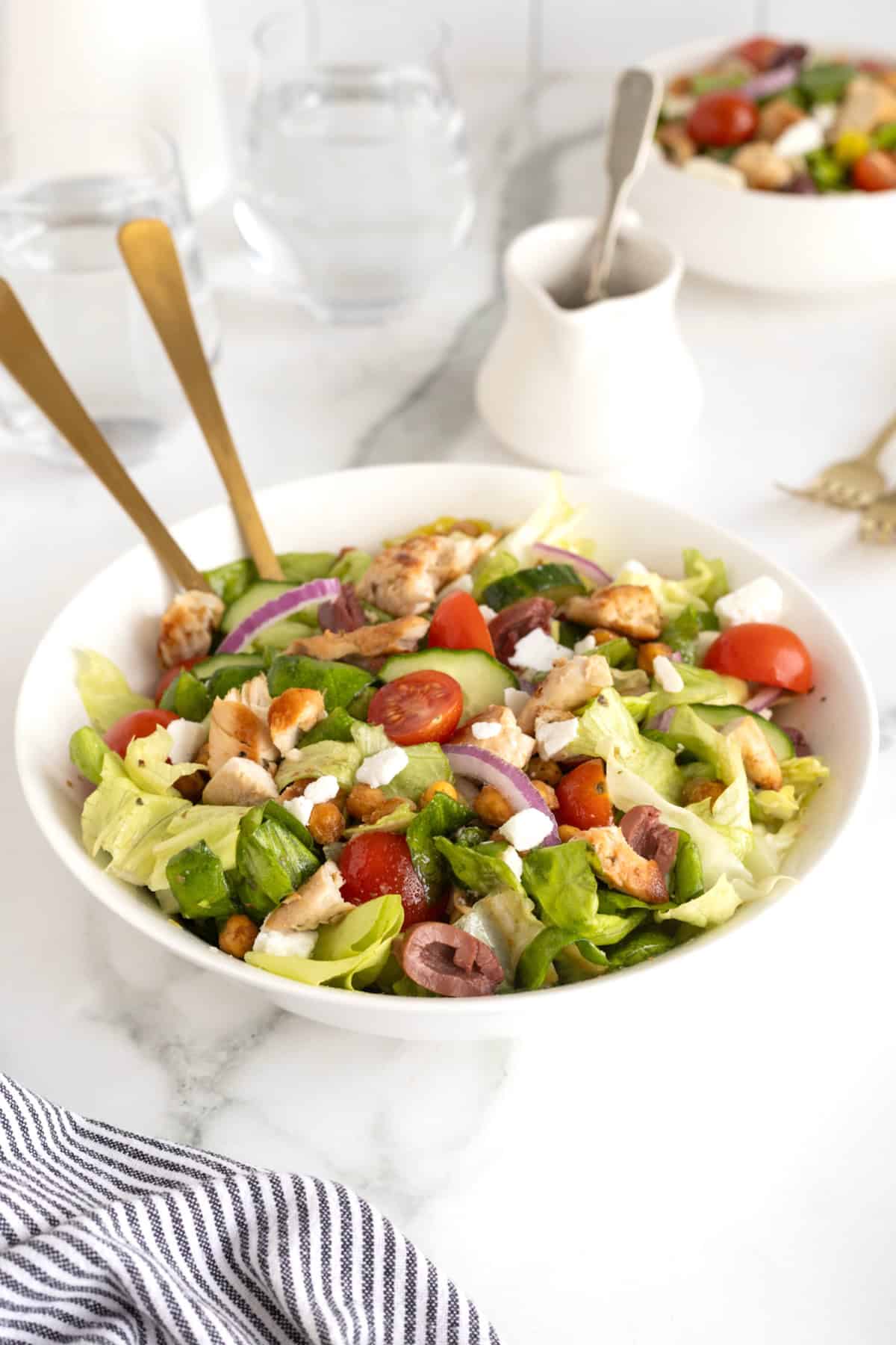 A large white serving bowl with greek salad topped with chicken, tomatoes, sliced greek olives, feta, and roasted chickpeas.