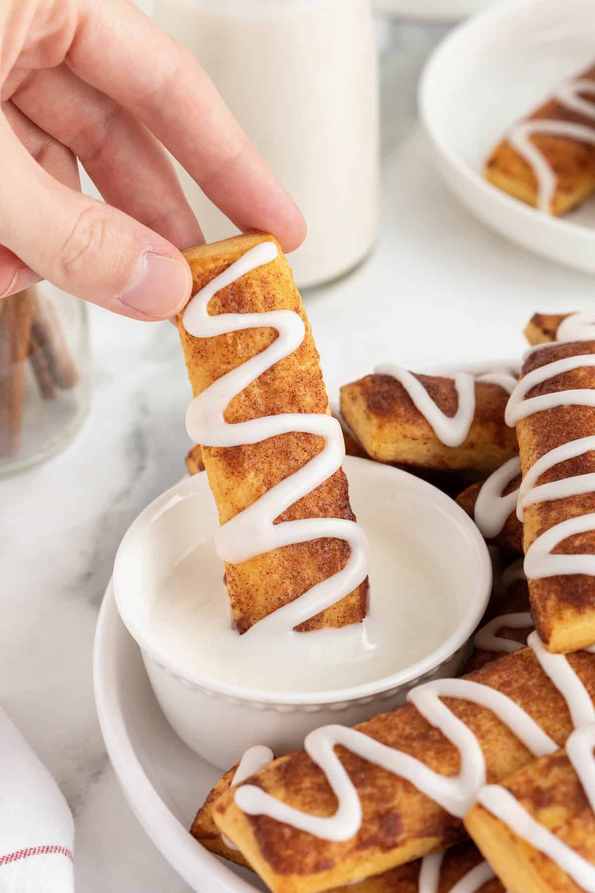 A hand dipping cinnamon roll stick into white icing.