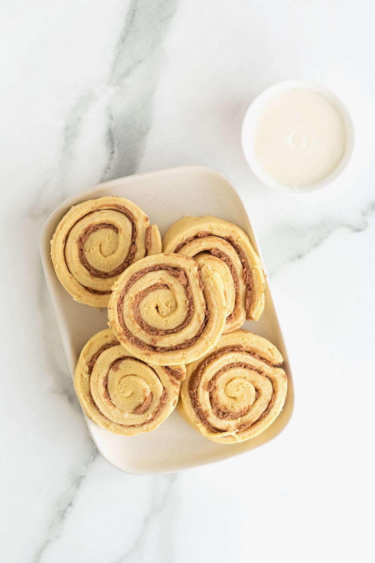 A white rectangular plate of canned cinnamon rolls next to a small white dish of icing on a white marble counter.