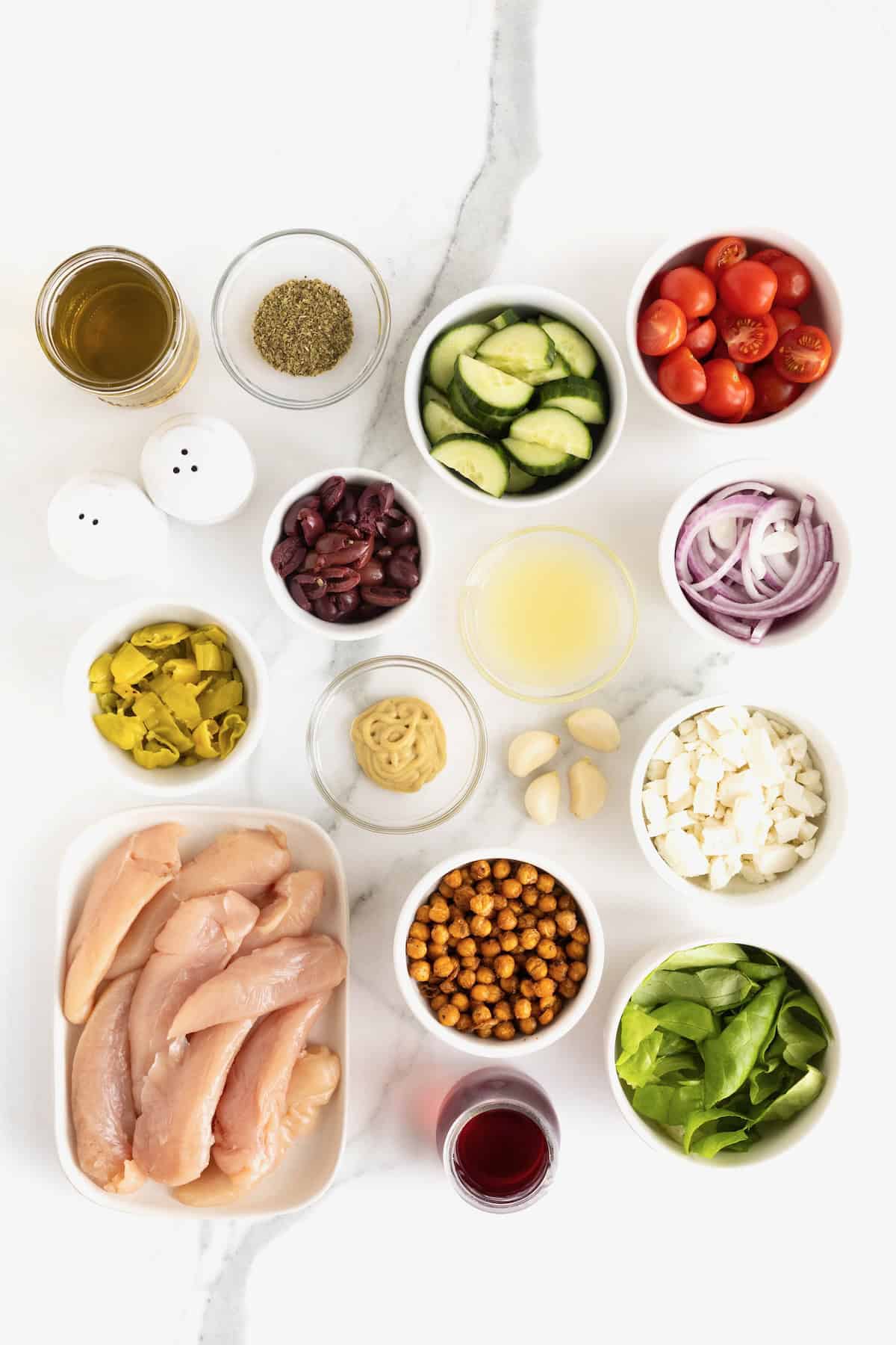 Ingredients to make Greek Salad with Marinated Chicken in small glass dishes on a white marble counter.