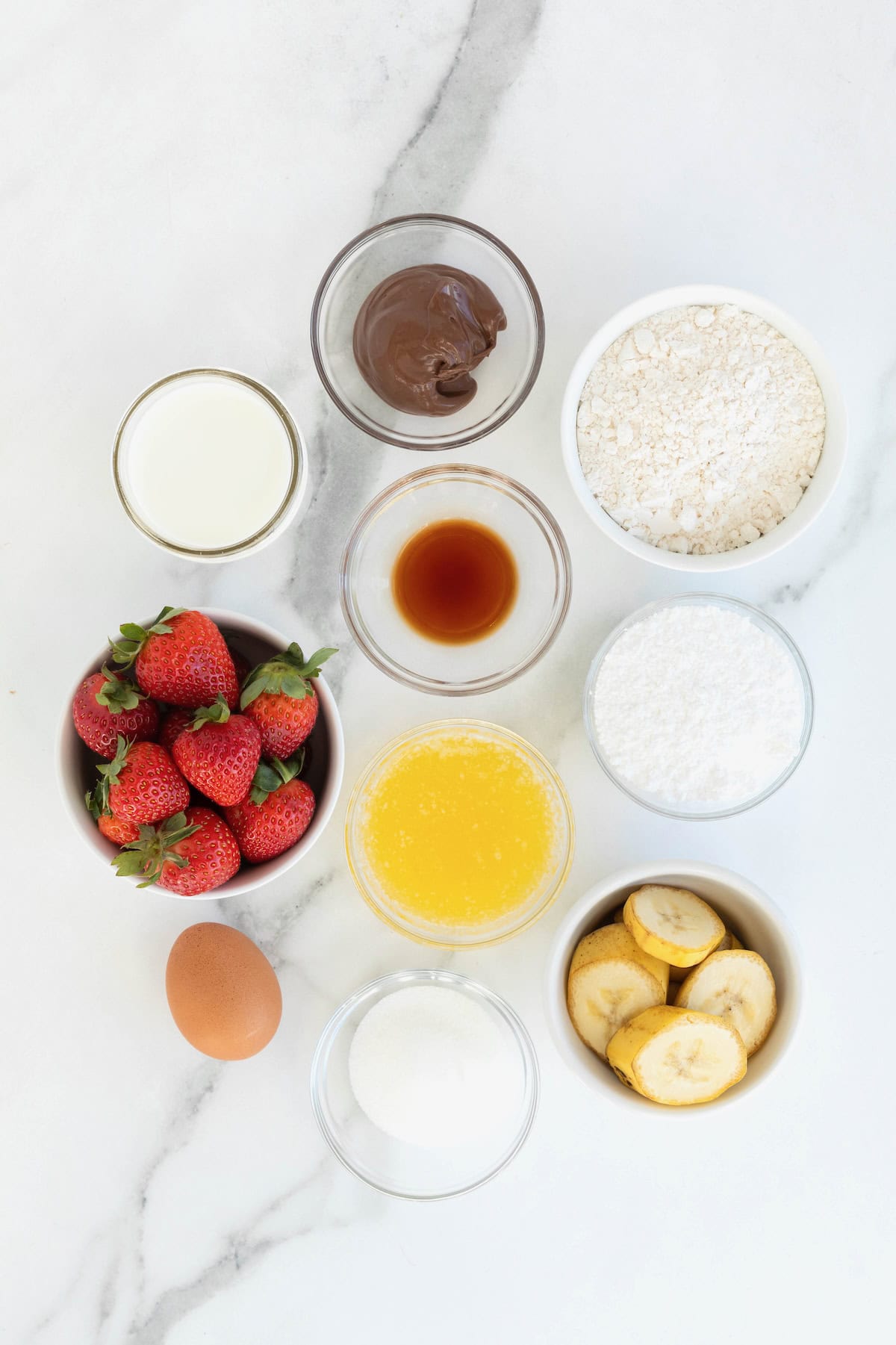 Ingredients to make sweet crepes in small glass dishes on a white marble counter.