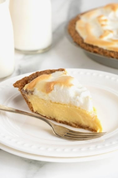 A slice of Lemon Meringue Pie next to a fork on a white dessert plate with beaded details.