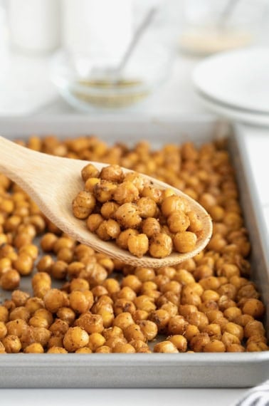 A wooden spoon of chickpeas in front of an aluminum baking sheet of roasted chickpeas.