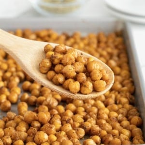 A wooden spoon of chickpeas in front of an aluminum baking sheet of roasted chickpeas.