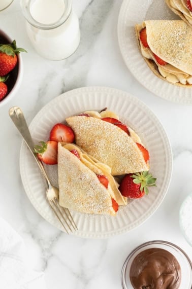 Two crepes on a white fluted plate with a fork. The crepes are filled with strawberry slices and banana slices and dusted with powdered sugar.