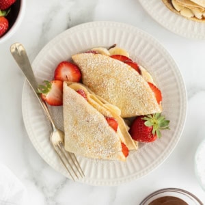 Two crepes on a white fluted plate with a fork. The crepes are filled with strawberry slices and banana slices and dusted with powdered sugar.